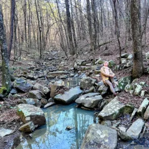 woman sitting on rocks by blue stream pool looking up at trees