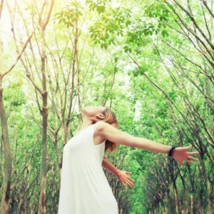 women in white dress stretching arms back in sunlit forest